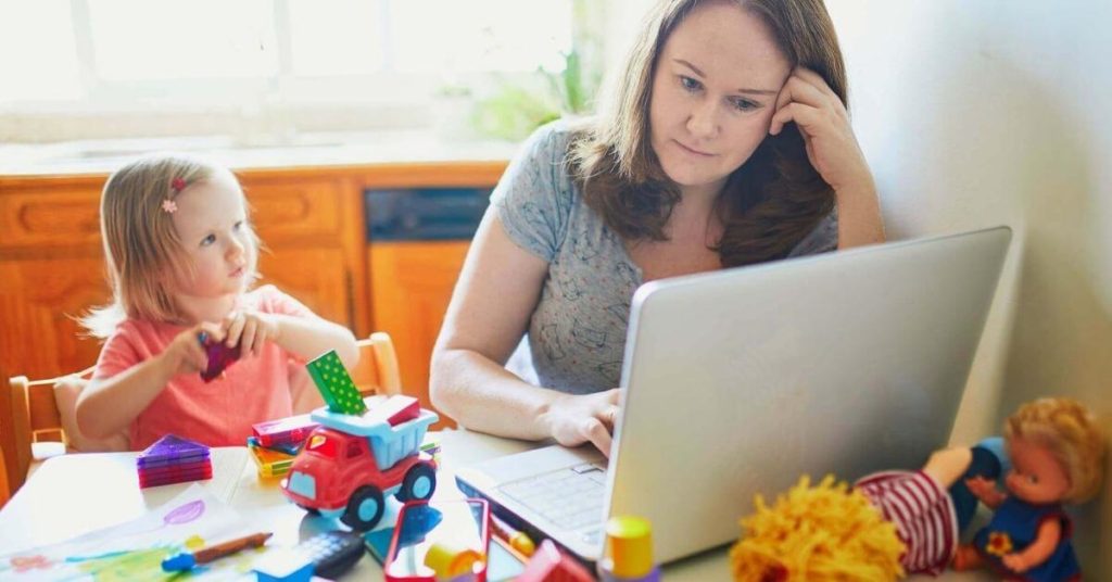 teacher mom working on a laptop with toddler playing with toys